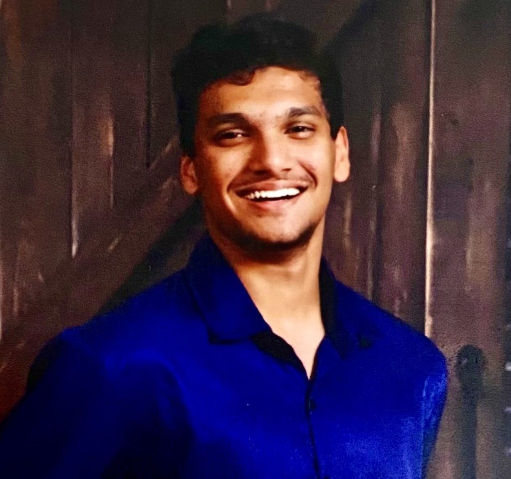 A man in a blue shirt smiles and stands against a dark wooden background.