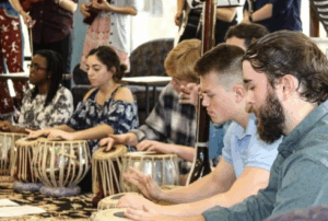 A group of people sit in a row playing tabla drums, focusing intently on their instruments during a musical performance or workshop.