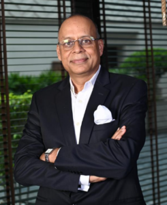 A man in a dark suit and glasses stands with arms crossed, smiling, in front of window blinds with greenery outside.