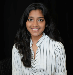 A woman with long dark hair wearing a white blouse with black stripes sits and smiles in front of a plain dark background.