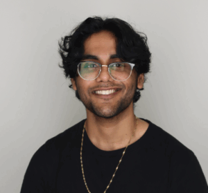 A young man with wavy dark hair, glasses, and a short beard smiles at the camera. He is wearing a black shirt and a gold chain necklace against a plain light background.