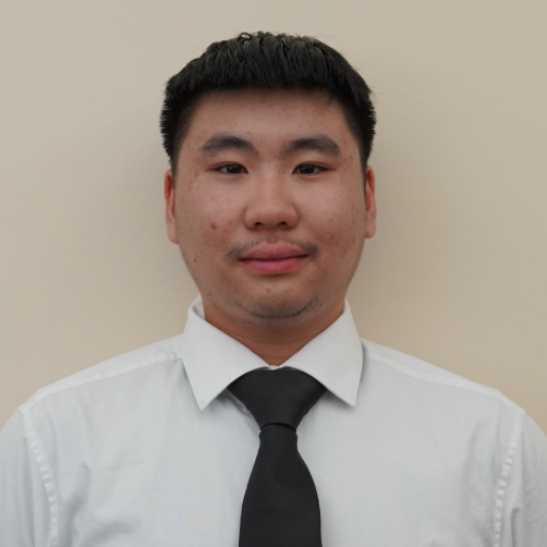 A man with short black hair wearing a white dress shirt and black tie stands against a plain beige background, looking at the camera.