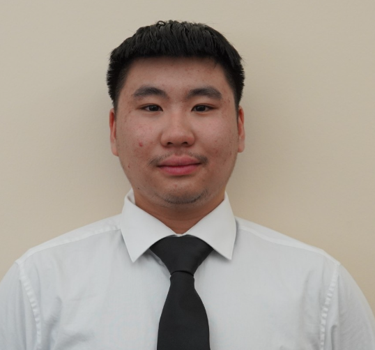 A man with short black hair wearing a white dress shirt and black tie stands against a plain beige background, looking at the camera.