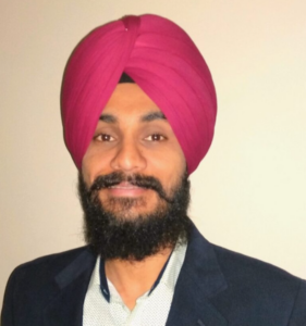 A man with a full beard wearing a bright pink turban, a light-colored shirt, and a dark blazer stands against a plain background.