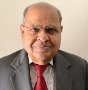 An older man wearing glasses, a grey suit, a checked shirt, and a red tie stands in front of a plain light-colored wall.