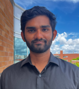 A man with short dark hair and a beard stands outside in front of a brick wall, wearing a black button-up shirt; the sky is blue with clouds.