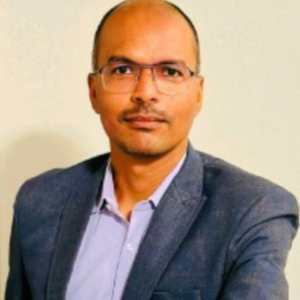 A man with glasses and a mustache, wearing a blue blazer and light-colored shirt, poses in front of a plain background.