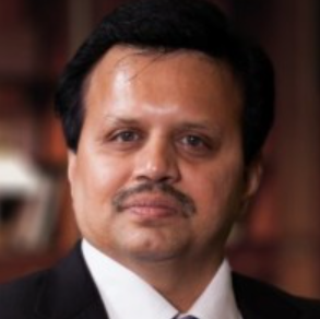 A man with dark hair and a mustache is wearing a dark suit, white shirt, and tie, posing for a formal portrait against a blurred background.