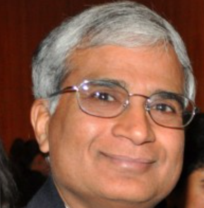 A middle-aged person with short gray hair, glasses, and a slight smile, wearing a dark suit, photographed against a blurred indoor background.