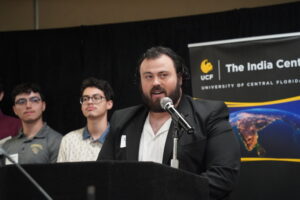A man in a suit speaks at a podium with a microphone, flanked by three men, with a UCF India Center banner in the background.