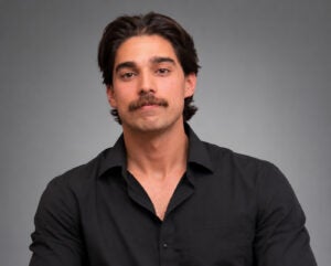 Man with dark hair and a mustache wearing a black buttoned shirt poses in front of a plain gray background.