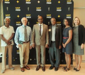 Six adults stand in a row, posing and smiling in front of a UCF Med backdrop. Four men and two women are dressed in business or business casual attire.