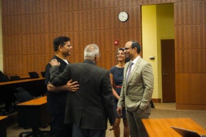Four people stand and talk in a conference room with wood-paneled walls, empty chairs, a clock, and a closed door in the background.