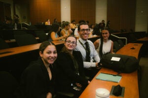 Four people sit at a table in a lecture hall, smiling at the camera. Other attendees are visible in the background.