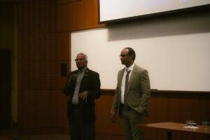 Two men in business attire stand at the front of a conference room, one speaking into a microphone while the other listens. A projection screen and a table with cups are visible behind them.
