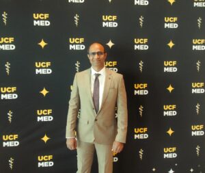 A man in a tan suit stands in front of a step-and-repeat banner featuring the UCF Med logo and medical symbols.