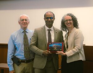 Three people stand together indoors; the person in the center, wearing a suit, holds a plaque, while the two others, wearing business attire, stand on each side and smile at the camera.