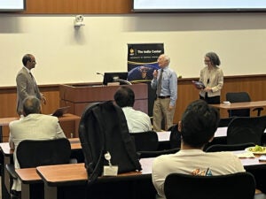 Three people stand at the front of a classroom near The India Center sign, while attendees watch from desks in the foreground.