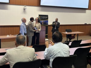 Several people in business attire stand and clap in a lecture hall as one person hands a plaque to another in front of a display reading The India Center.