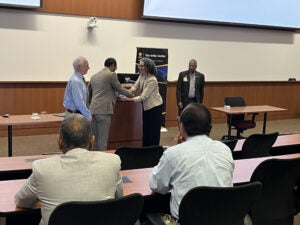 Four people stand and shake hands at the front of a lecture hall while three seated attendees observe. A banner for The India Center is visible in the background.