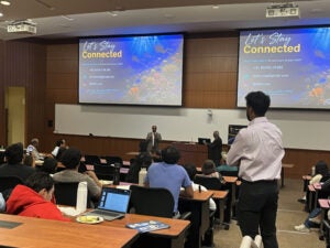 Two speakers address an audience in a lecture hall. Slides on the screen display contact information and the message Let's Stay Connected. Audience members are seated and attentive.