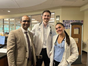 Three people, two in white medical coats with stethoscopes and one in a suit, stand together and smile in a hospital or medical office setting.