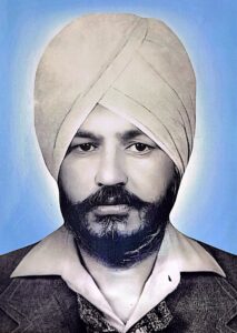 A man wearing a turban, collared shirt, and blazer, with a beard and mustache, poses for a formal portrait against a blue background.