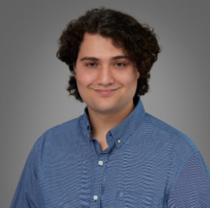 A young person with medium-length curly hair, wearing a blue button-up shirt, poses against a plain gray background.