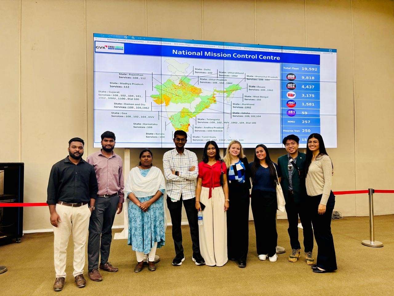 A group of nine people stand in front of a large display screen showing a map and statistics at the National Mission Control Centre.