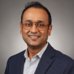 A man wearing glasses, a checkered shirt, and a navy blazer is smiling at the camera against a plain gray background.