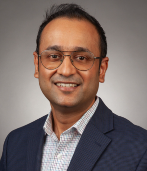 A man wearing glasses, a checkered shirt, and a navy blazer is smiling at the camera against a plain gray background.