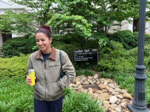 A person stands outside near a sign for Candler Hall, holding a canned drink and smiling, with greenery and a lamppost in the background.