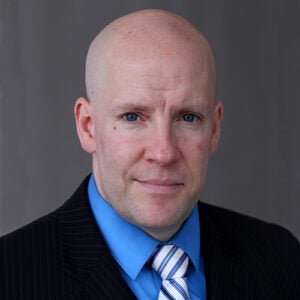 A bald man wearing a dark pinstripe suit, blue shirt, and striped tie looks at the camera against a plain gray background.
