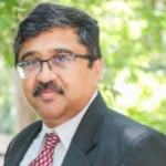 Man with glasses and mustache wearing a dark suit, white shirt, and red patterned tie, standing outdoors with greenery in the background.