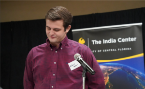 A man in a red checkered shirt stands at a microphone, looking down. Behind him is a banner for The India Center at the University of Central Florida.