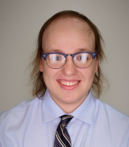 A smiling person with glasses, light brown hair, and a receding hairline, wearing a light blue dress shirt and striped tie, poses in front of a plain background.