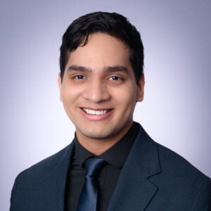 Young man with short dark hair wearing a dark suit, black shirt, and navy tie, smiling in front of a plain light purple background.