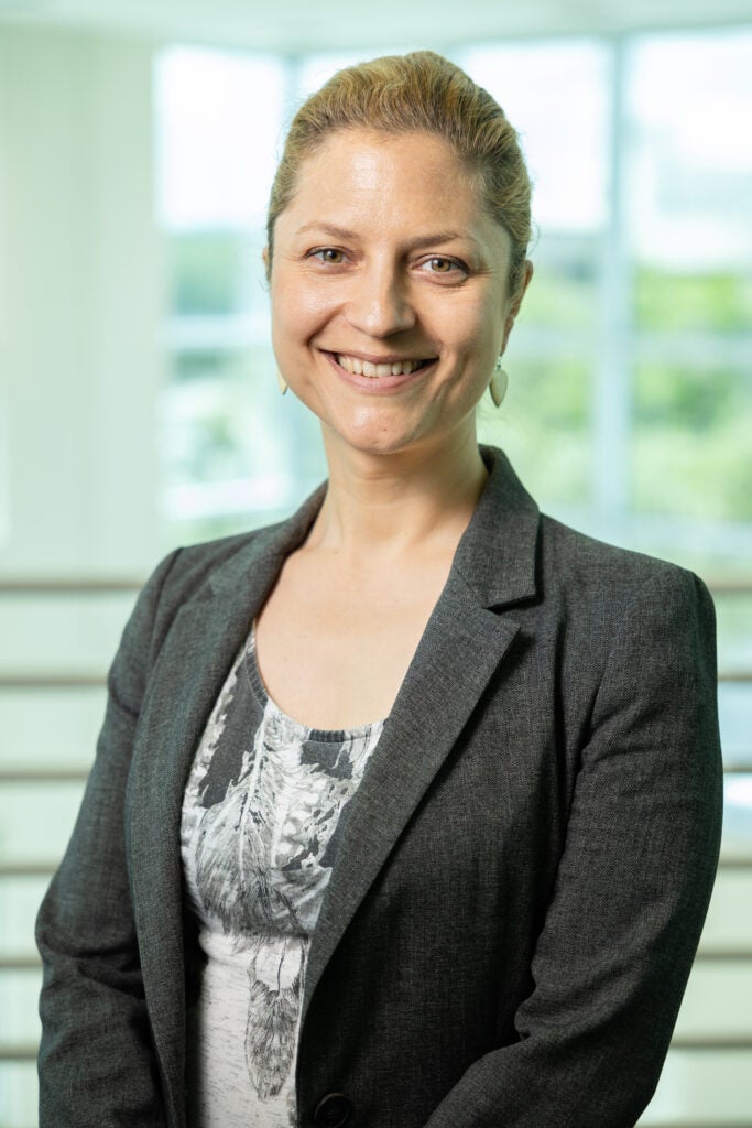 A woman with blonde hair pulled back, wearing a gray blazer over a patterned top, smiling at the camera.