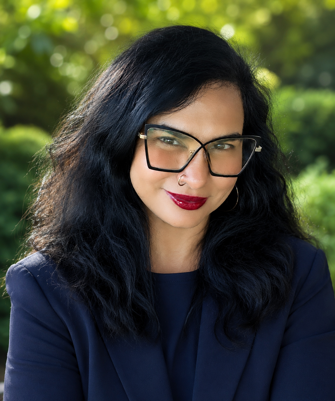 A woman with dark hair, glasses, and dark red lipstick poses outdoors.