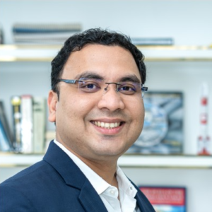 A headshot of a smiling man wearing glasses and a navy blue blazer.
