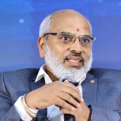 Sudheer N. Kumar, wearing a suit and conference badge, speaks into a microphone at an event.
